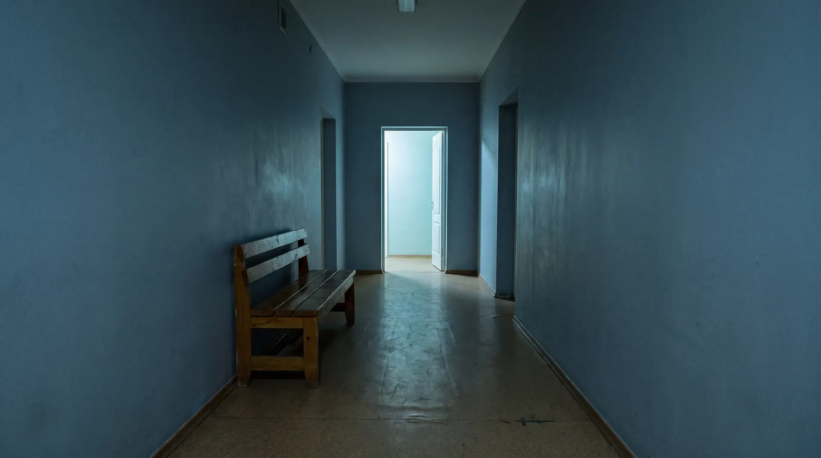 An empty blue-grey hallway before dawn with a wooden bench and one lit doorway at the end