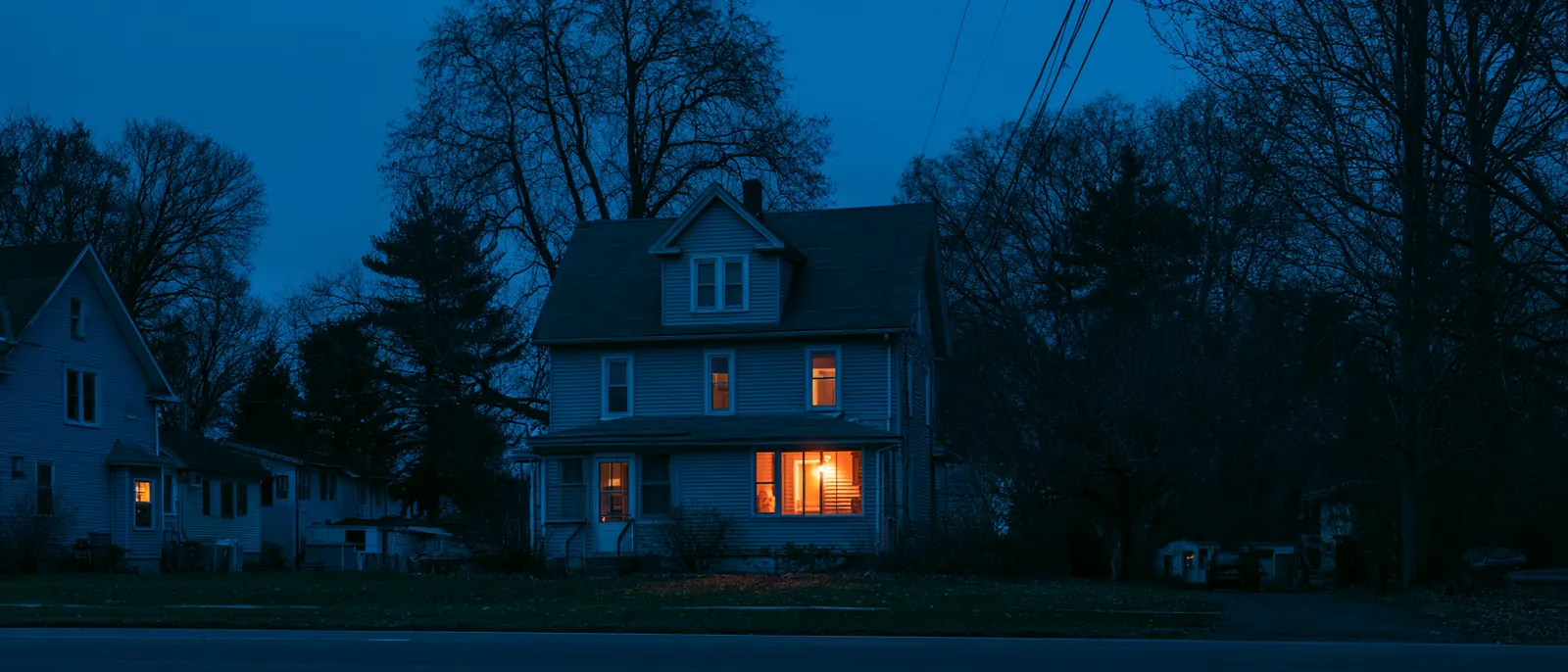 A two-story house at night with one downstairs window glowing warm, bare trees around it